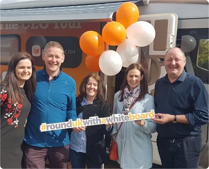 Five OnePageCRM customers holding a #RoundUKwithaWhiteboard sign in front of the orange CEO Tour camper van, with orange and white balloons above them